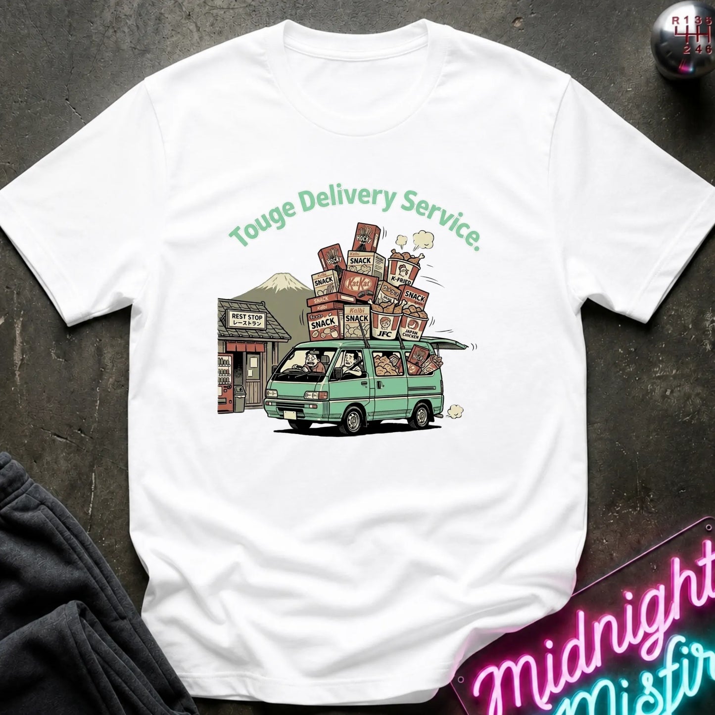 A white  short-sleeve t-shirt with a graphic print of a green van carrying stacked cardboard boxes under the text 'Touge Delivery Service.' The shirt is laid flat on a dark textured surface with a neon sign and dark fabric partially visible at the edges.