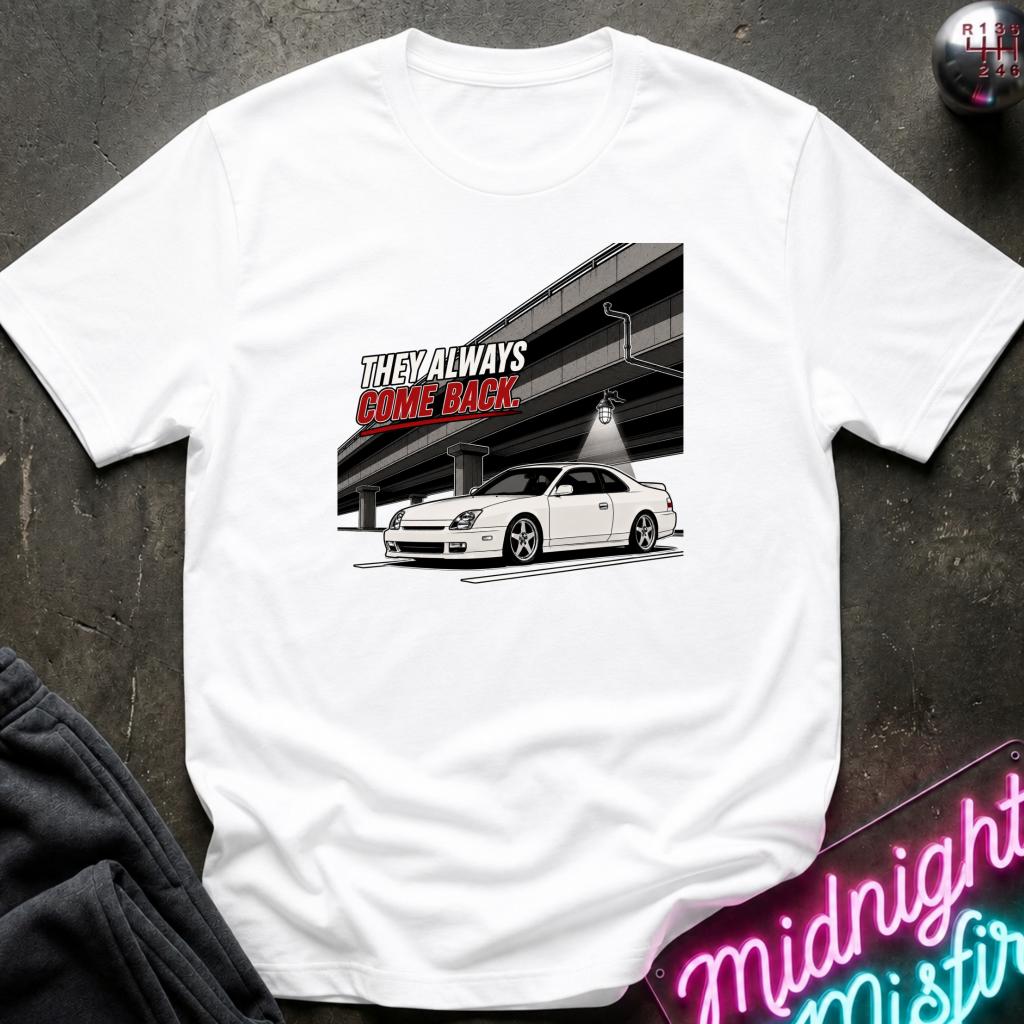 A white short-sleeve graphic t-shirt laid flat on a dark textured surface. The graphic features a white sports car under a bridge with the text 'THE WAY ALWAYS CAME BACK' in red and black.