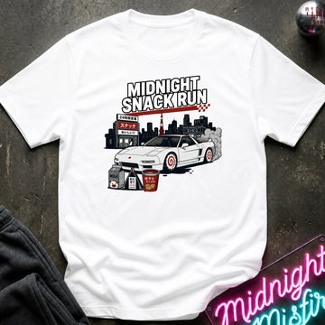 A white short-sleeve graphic t-shirt laid flat on a dark textured surface. The graphic features the text 'MIDNIGHT SNACKRUN' above an illustration of a white sports car, a city skyline, and snack boxes.