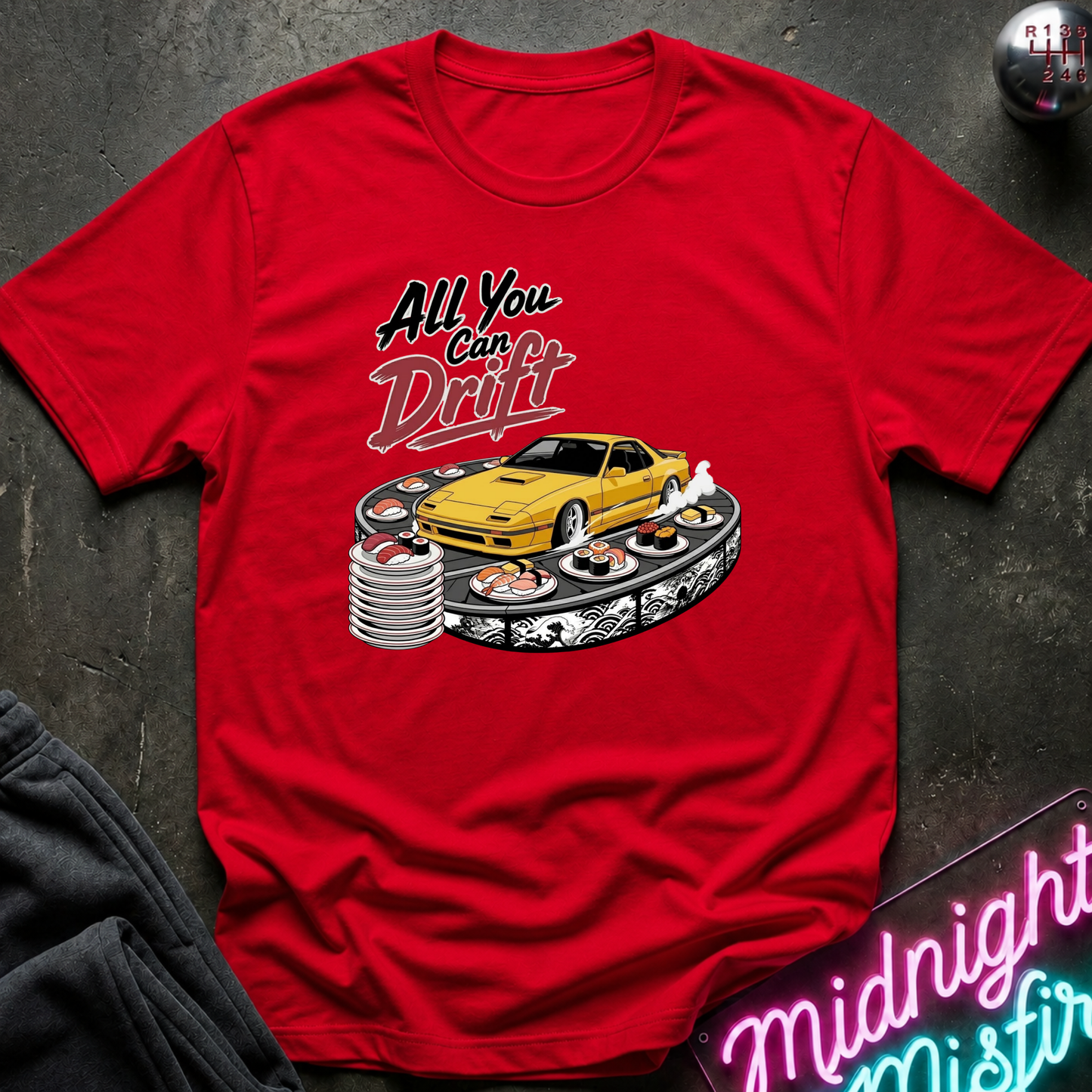 A red short-sleeve graphic T-shirt laid flat on a dark textured surface. The shirt features a printed design of a yellow sports car drifting on a track with the text 'All You Can Drift' above it.