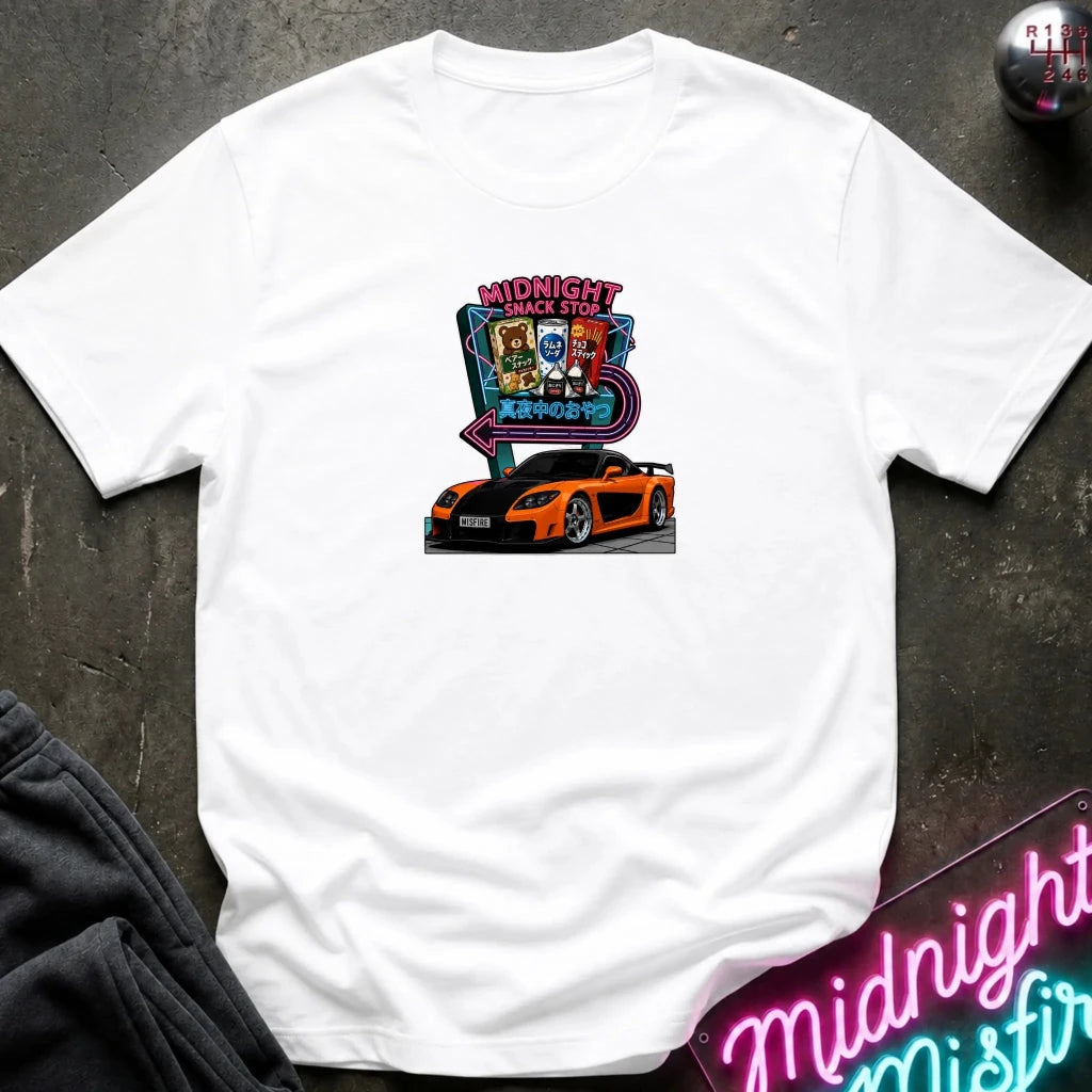 A white short-sleeve t-shirt laid flat on a dark textured surface, featuring a colorful graphic print of an orange sports car in front of a neon sign that reads 'Midnight Snack Stop'.