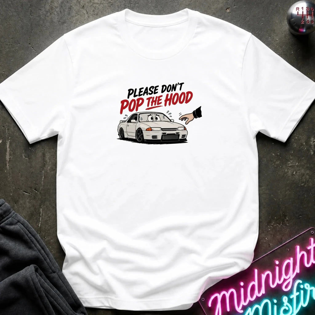 A white short-sleeve t-shirt laid flat on a dark textured surface, featuring a graphic of a white sports car with the text 'PLEASE DON'T POP THE HOOD' in black and red. A neon sign and dark clothing are partially visible as props.