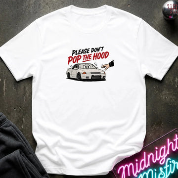 A white short-sleeve t-shirt laid flat on a dark textured surface, featuring a graphic of a white sports car with the text 'PLEASE DON'T POP THE HOOD' in black and red. A neon sign and dark clothing are partially visible as props.