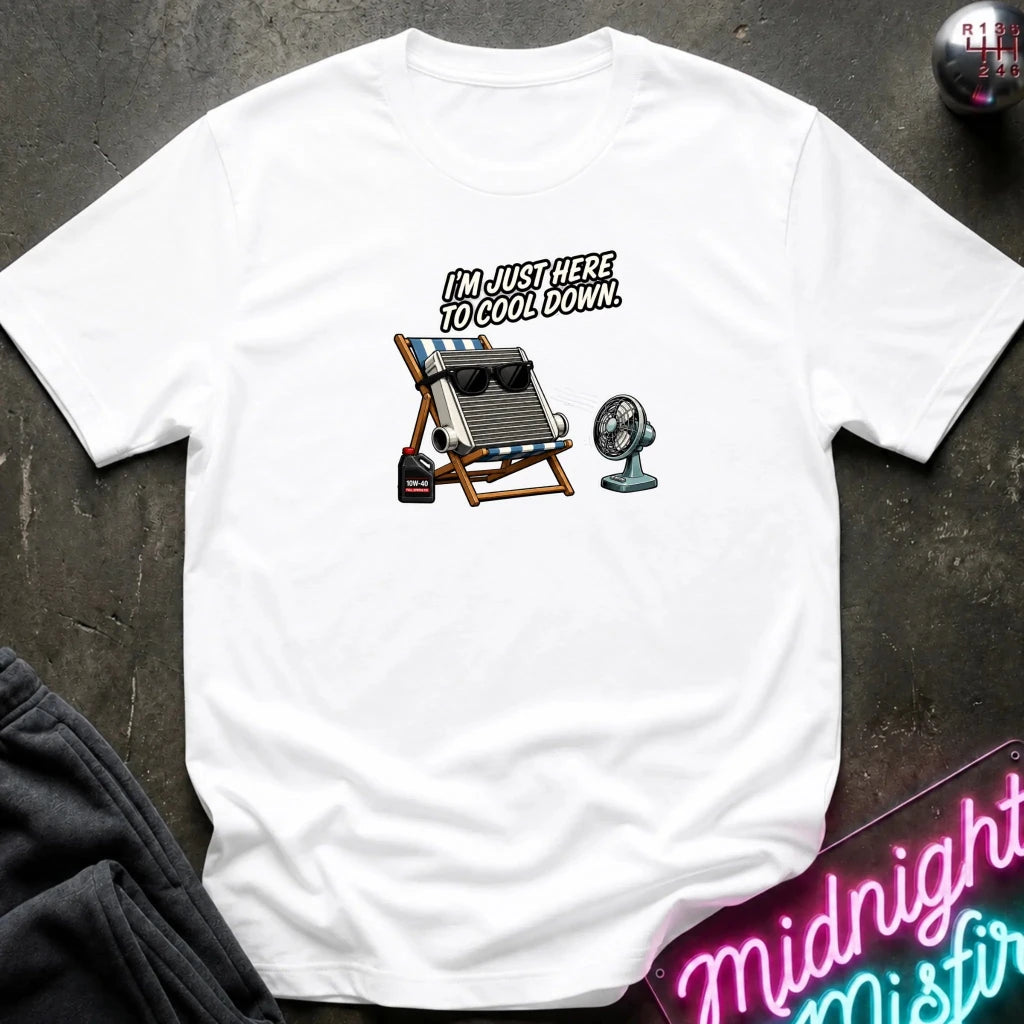 A white short-sleeve graphic t-shirt laid flat on a dark textured surface. The graphic features with the text 'I'M JUST HERE TO COOL DOWN!' above it.