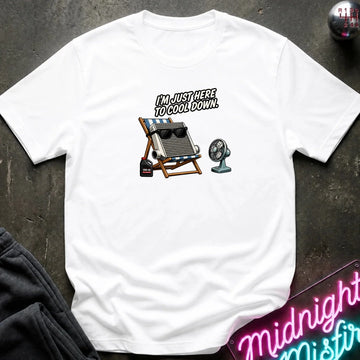 A white short-sleeve graphic t-shirt laid flat on a dark textured surface. The graphic features with the text 'I'M JUST HERE TO COOL DOWN!' above it.