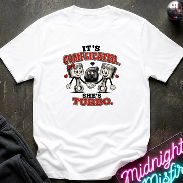 A white short-sleeve graphic T-shirt laid flat on a dark textured surface. The shirt features a cartoon chest print with two pistons, a black turbo, and the text 'IT'S COMPLICATED.