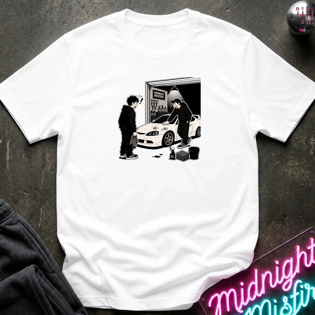 A white short-sleeve t-shirt laid flat on a dark textured surface, featuring a black and white graphic print of a person standing next to a white sports car in front of a garage door. 