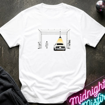A white short-sleeve t-shirt laid flat on a dark textured surface, featuring a graphic print of a car under a hanging lamp inside a garage structure. 
