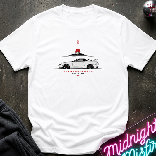 A white short-sleeve graphic T-shirt laid flat on a dark textured surface. The shirt features a printed illustration of a white sports car with a red sun and mountain graphic above it, along with small red and black text.