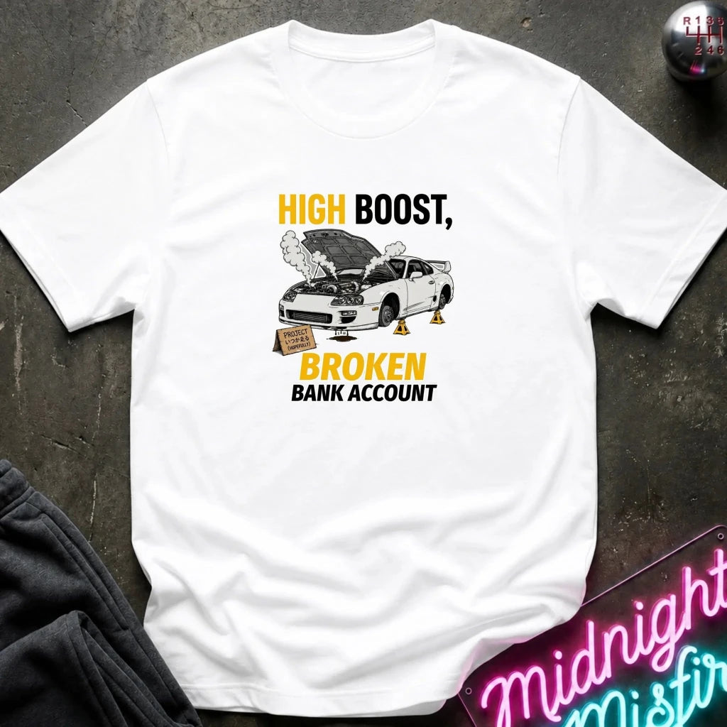 A white short-sleeve graphic T-shirt laid flat on a dark textured surface. The shirt features a printed design with the text 'HIGH BOOST, BROKEN BANK ACCOUNT' in black and yellow, alongside an illustration of a white car with its hood open and oil cans.