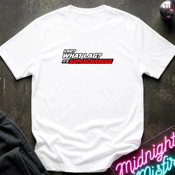 A white  short-sleeve t-shirt with a graphic text print reading 'LAG? WHAT LAG? IT'S SUPERCHARGED' in white, black, and red, laid flat on a dark textured surface with a gray sweatshirt and a neon sign partially visible as props.