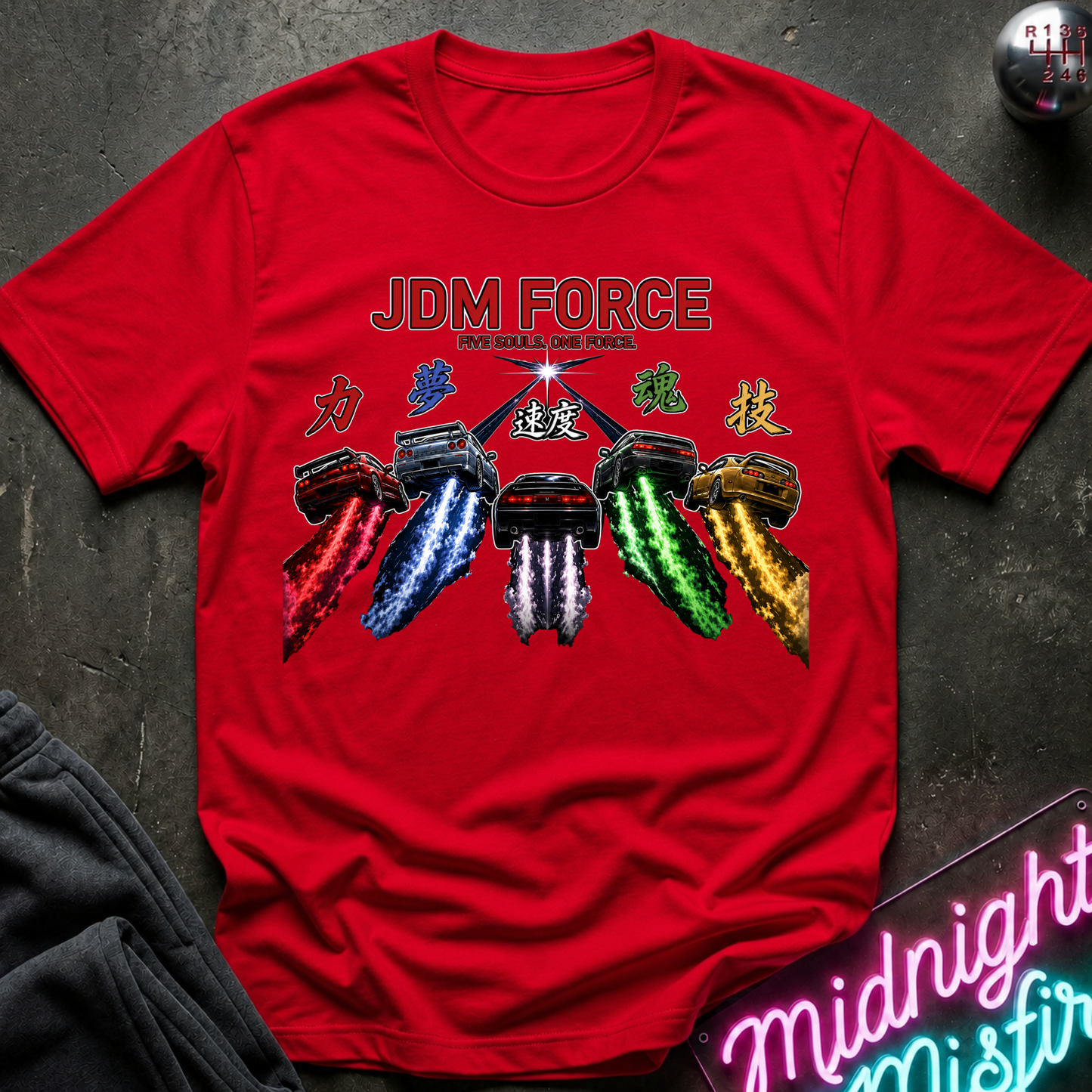 A bright red short-sleeve t-shirt laid flat on a dark textured surface. The shirt features a graphic print with the text 'JDM FORCE' and illustration of cars with colorful exhaust trails in red, blue, white, and green, with green Japanese characters.