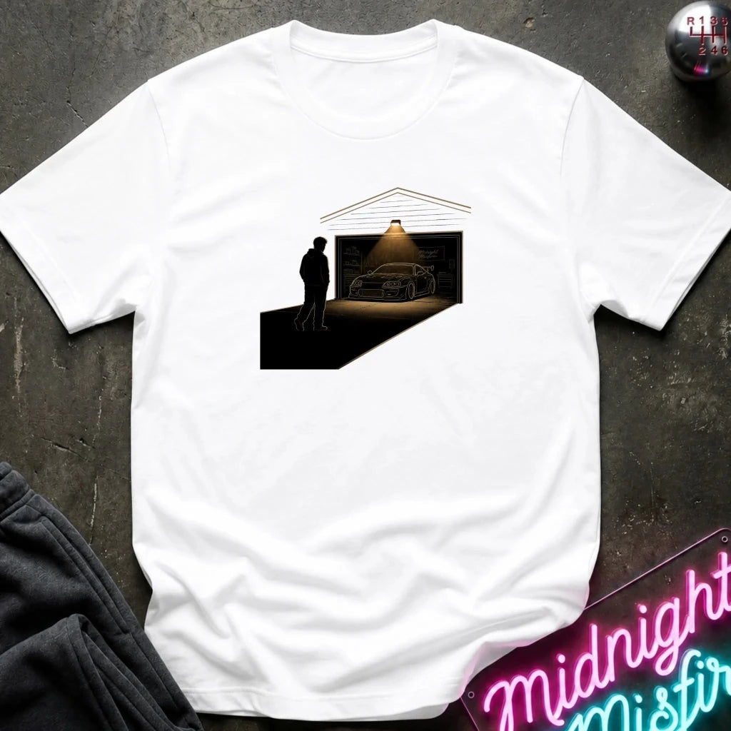 A white short-sleeve t-shirt laid flat on a dark textured surface, featuring a central graphic print of a silhouette standing in front of a garage with a car inside under a spotlight.