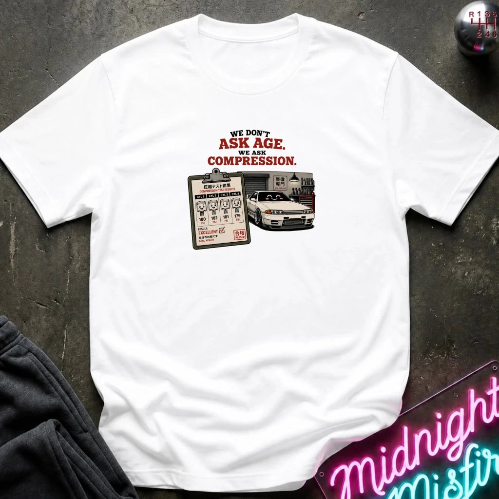 A white short-sleeve graphic t-shirt laid flat on a dark textured surface. The graphic features the text 'WE DON'T ASK AGE, WE ASK COMPRESSION.