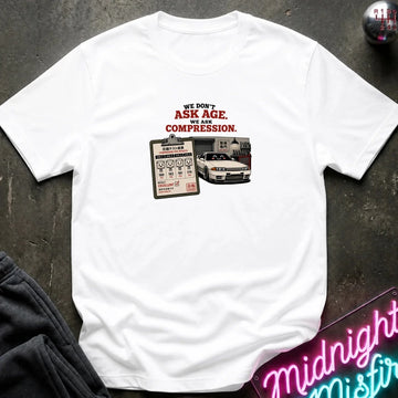 A white short-sleeve graphic t-shirt laid flat on a dark textured surface. The graphic features the text 'WE DON'T ASK AGE, WE ASK COMPRESSION.