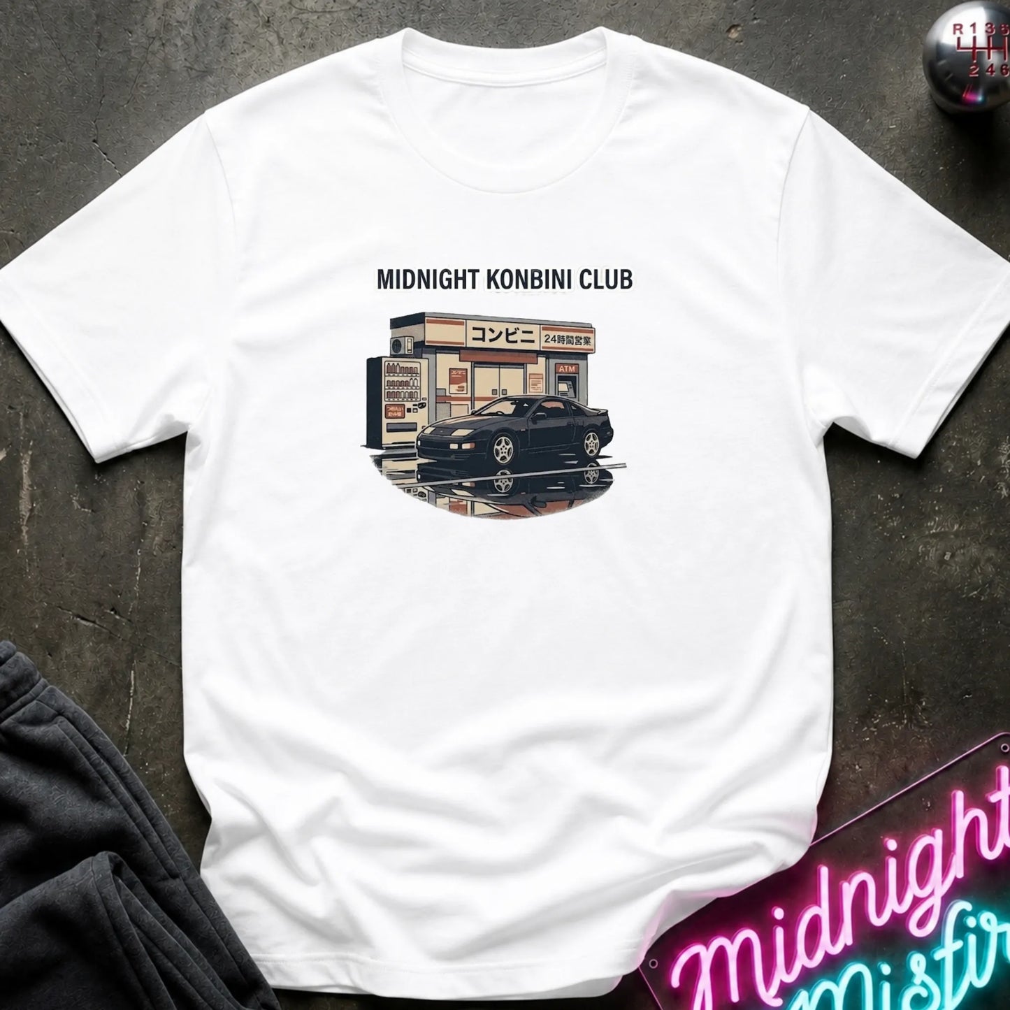 A white short-sleeve t-shirt laid flat on a dark textured surface, featuring a graphic print of a black sports car parked in front of a convenience store with the text 'MIDNIGHT KONBINI CLUB' above it.