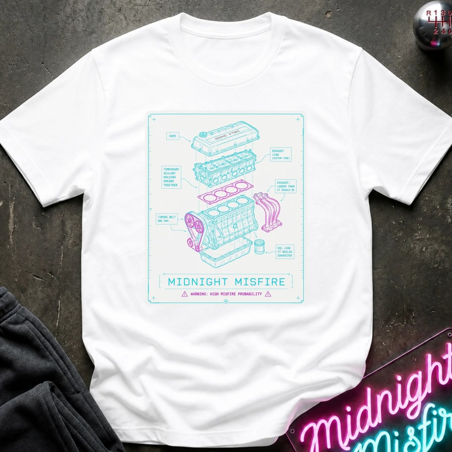 A white short-sleeve t-shirt laid flat on a dark textured surface, featuring a light blue and pink technical schematic graphic of an engine labeled 'MIDNIGHT MISFIRE'.