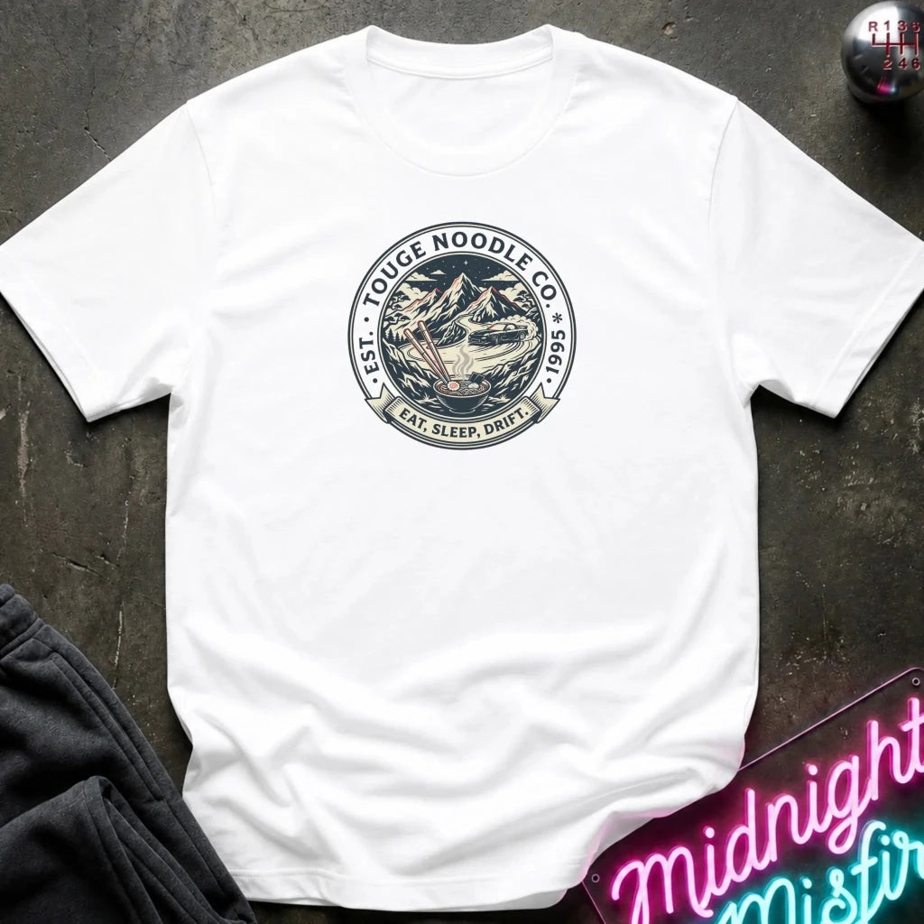 A white short-sleeve crewneck t-shirt laid flat on a dark textured surface. The center of the shirt features a circular graphic print with a noodle bowl illustration and the text 'TOUGE NOODLE CO' 