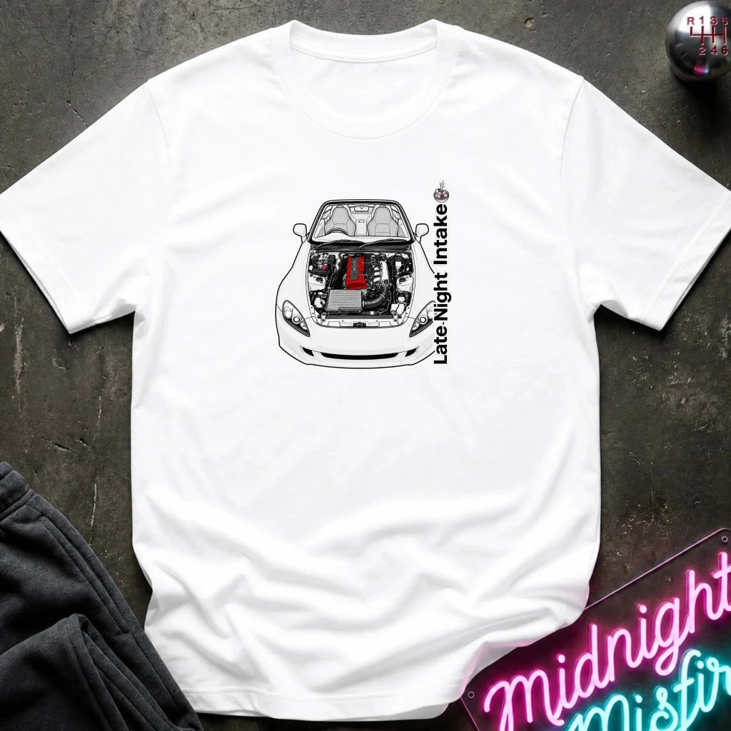 A white short-sleeve t-shirt laid flat on a dark textured surface, featuring a graphic of a white sports car with its hood open, revealing a red engine component, alongside the vertical text 'Late Night Intake'.