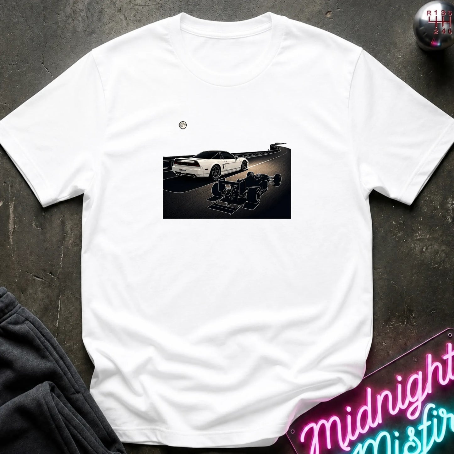 A white short-sleeve crewneck t-shirt laid flat on a dark textured surface, featuring a graphic print of a white sports car and a shadow car on a night road scene. The shirt is styled with dark gray sweatpants, a silver ball, and a neon sign in the corners.
