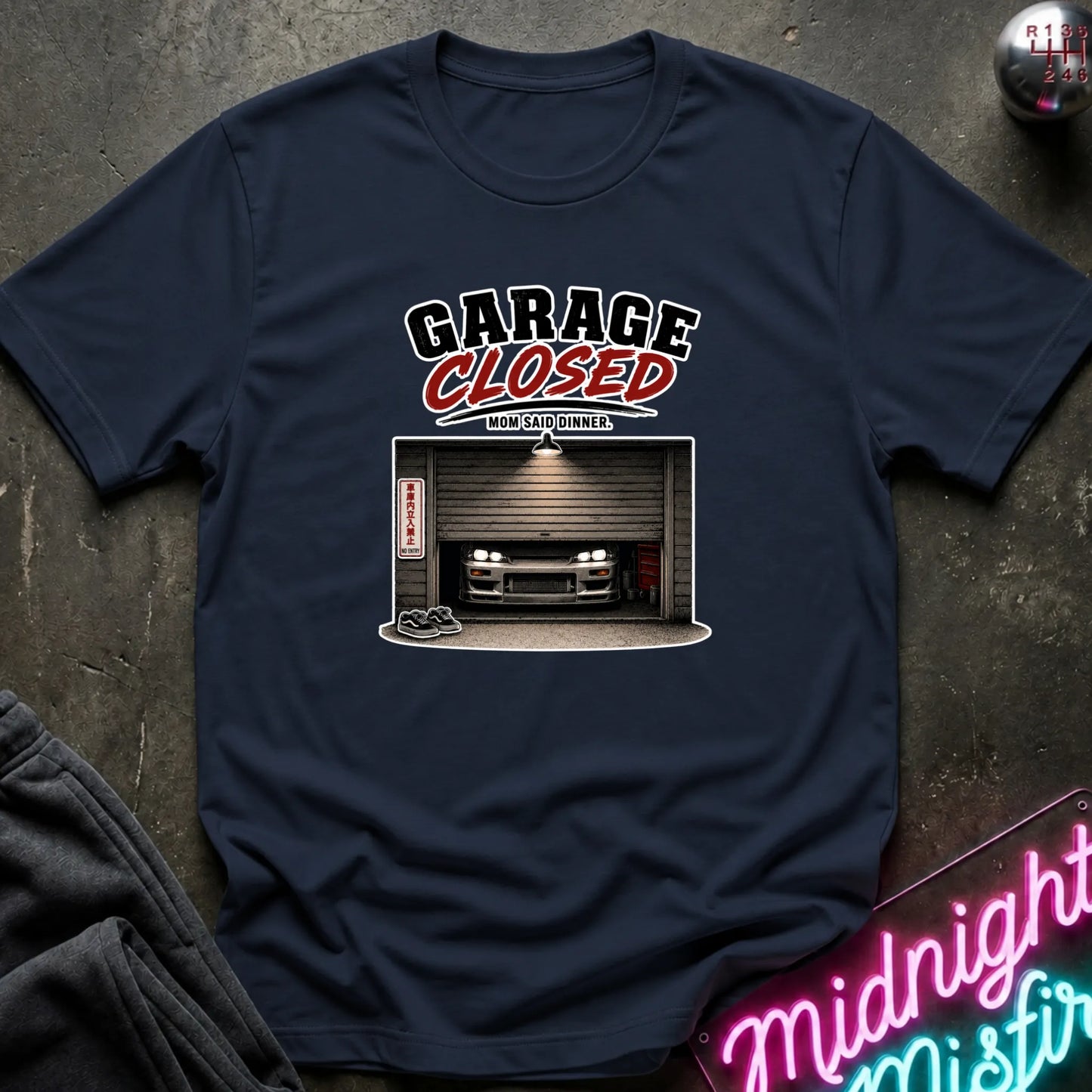 A navy blue graphic t-shirt laid flat on a dark textured surface. The shirt features a graphic print with the text 'GARAGE CLOSED' in white and red, above an illustration of a car parked inside a garage.