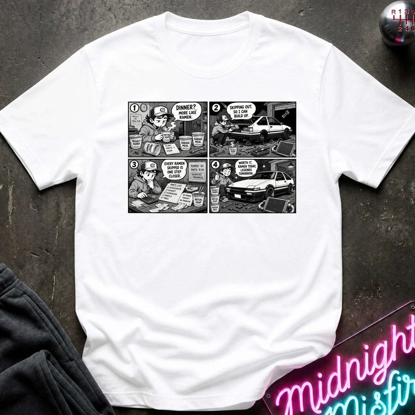 A white short-sleeve crewneck t-shirt featuring a black and white comic strip graphic on the chest, styled in a flat-lay on a dark textured surface with a neon sign and dark pants partially visible.