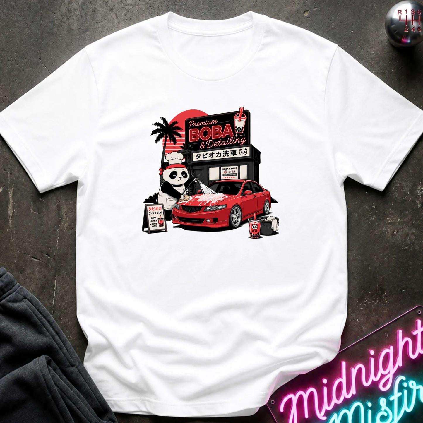 A white short-sleeve t-shirt laid flat on a dark textured surface, featuring a colorful graphic print of a panda mascot, a red sports car, and a Japanese-style detailing shop sign.