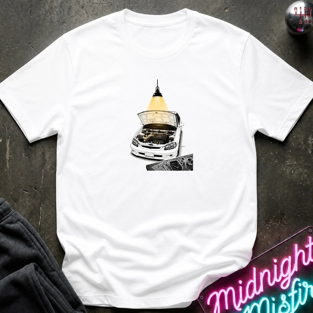 A white short-sleeve t-shirt laid flat on a dark textured surface, featuring a graphic print of a white car with its hood open under a glowing yellow lamp. 