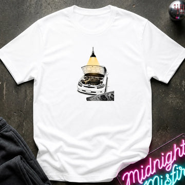 A white short-sleeve t-shirt laid flat on a dark textured surface, featuring a graphic print of a white car with its hood open under a glowing yellow lamp. 