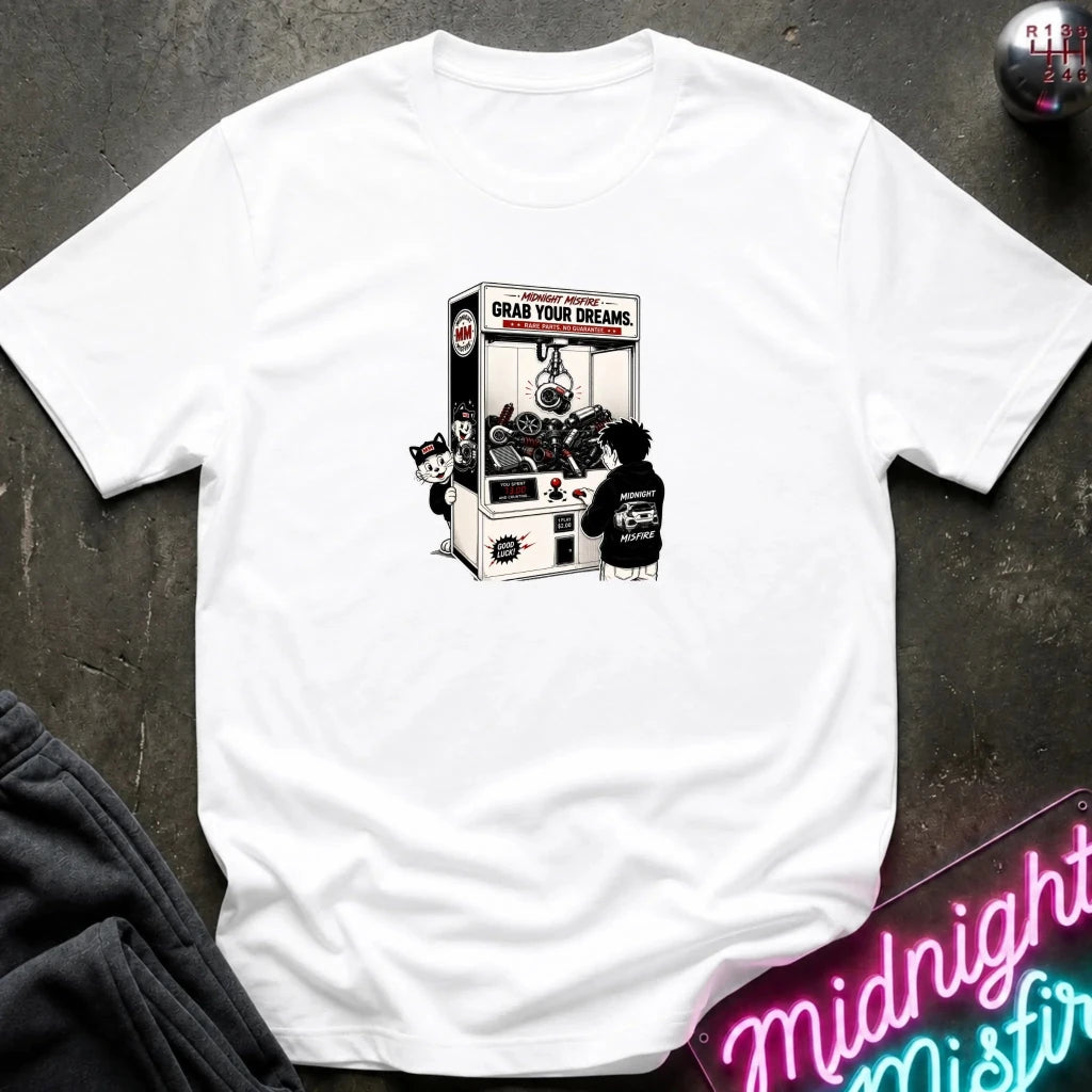 A white short-sleeve t-shirt laid flat on a dark textured surface, featuring a black and white graphic print of a claw machine with the text 'GRAB YOUR DREAMS'. 