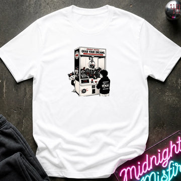 A white short-sleeve t-shirt laid flat on a dark textured surface, featuring a black and white graphic print of a claw machine with the text 'GRAB YOUR DREAMS'. 