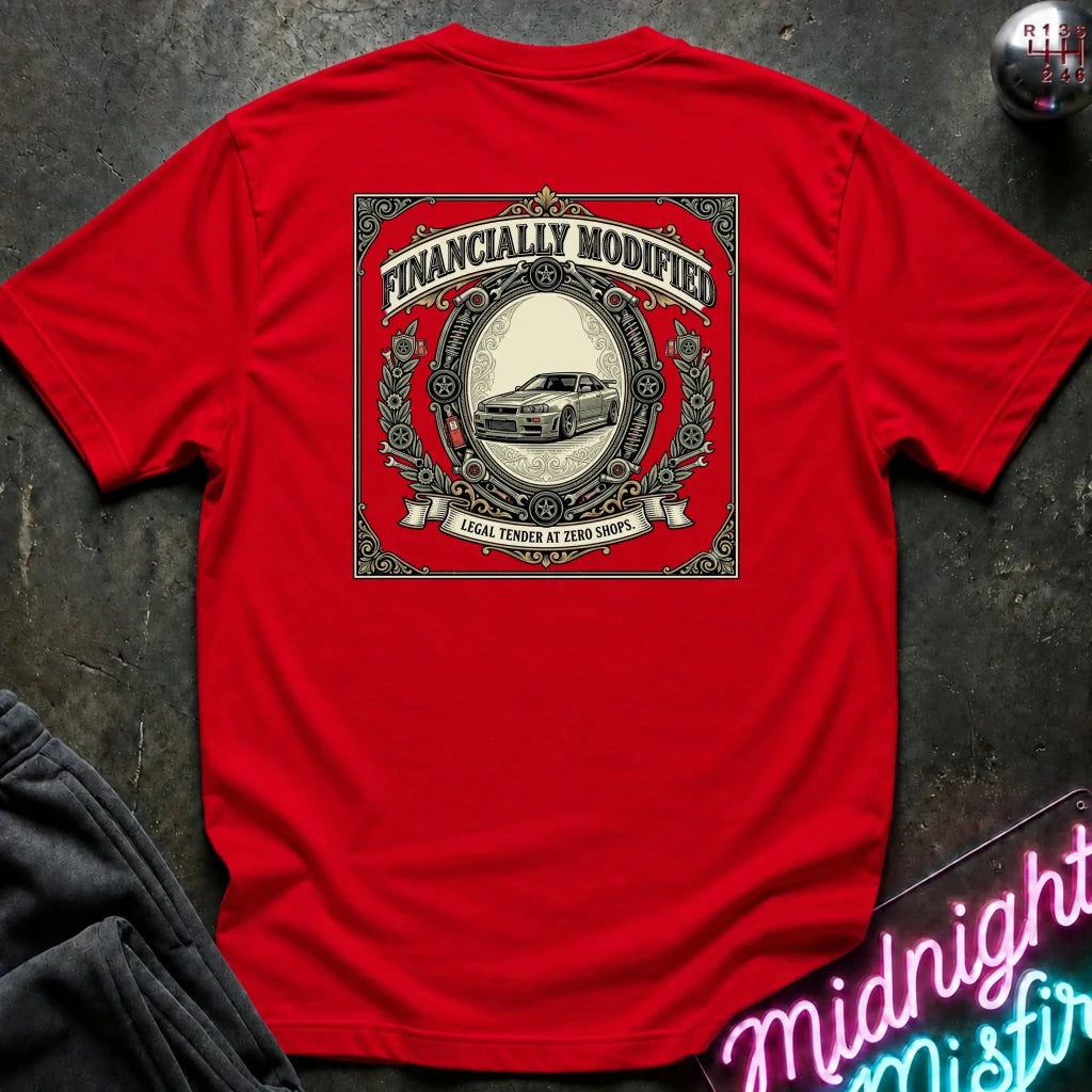 A bright red short-sleeve t-shirt laid flat on a dark textured surface, featuring a large vintage-style graphic print on the back with the words 'FINANCALLY MODIFIED' and an illustration of a car. A neon sign and dark clothing are partially visible around the edges as styling props.