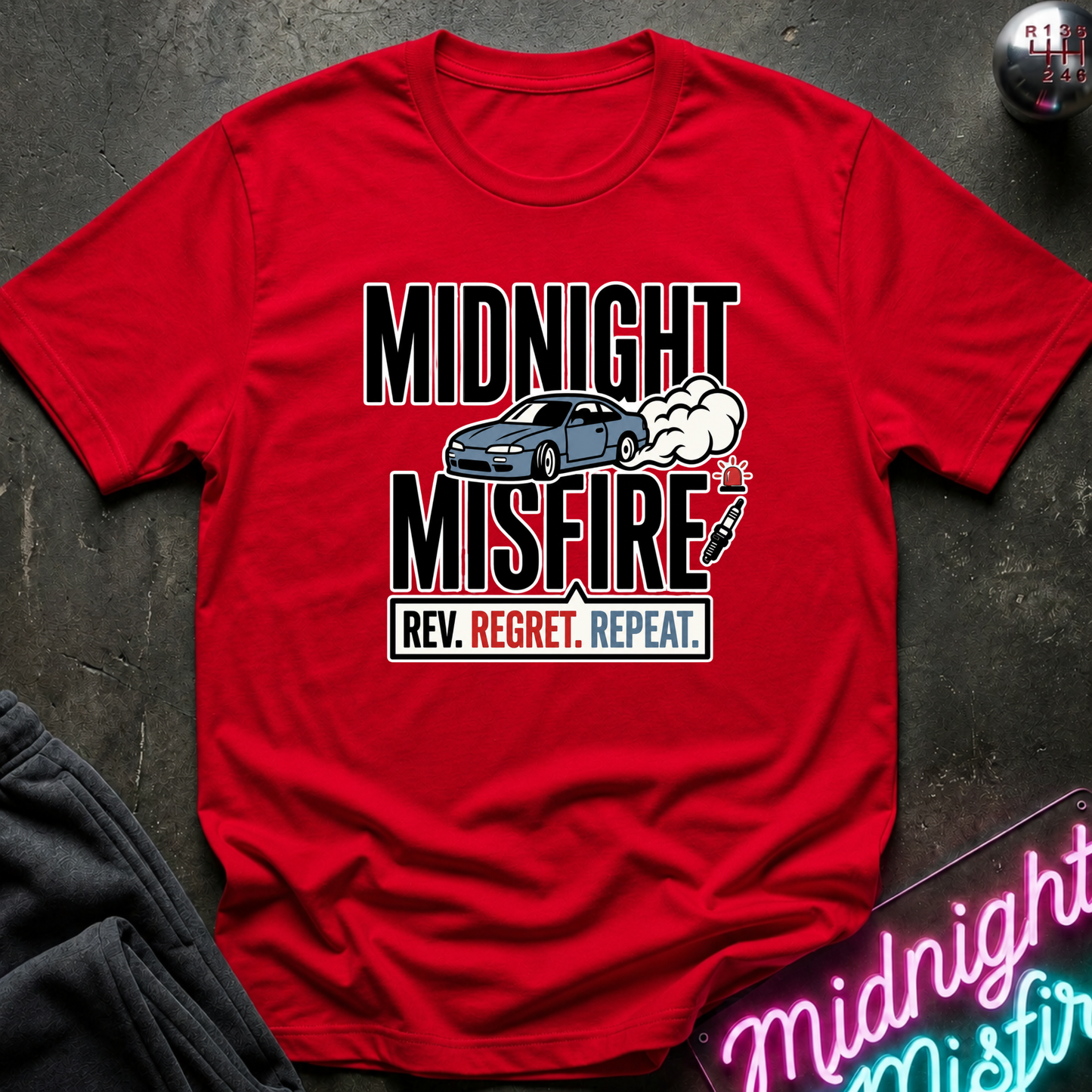 A red short-sleeve graphic T-shirt laid flat on a dark textured surface. The shirt features a black, white, and gray print that reads 'MIDNIGHT MISFIRE.