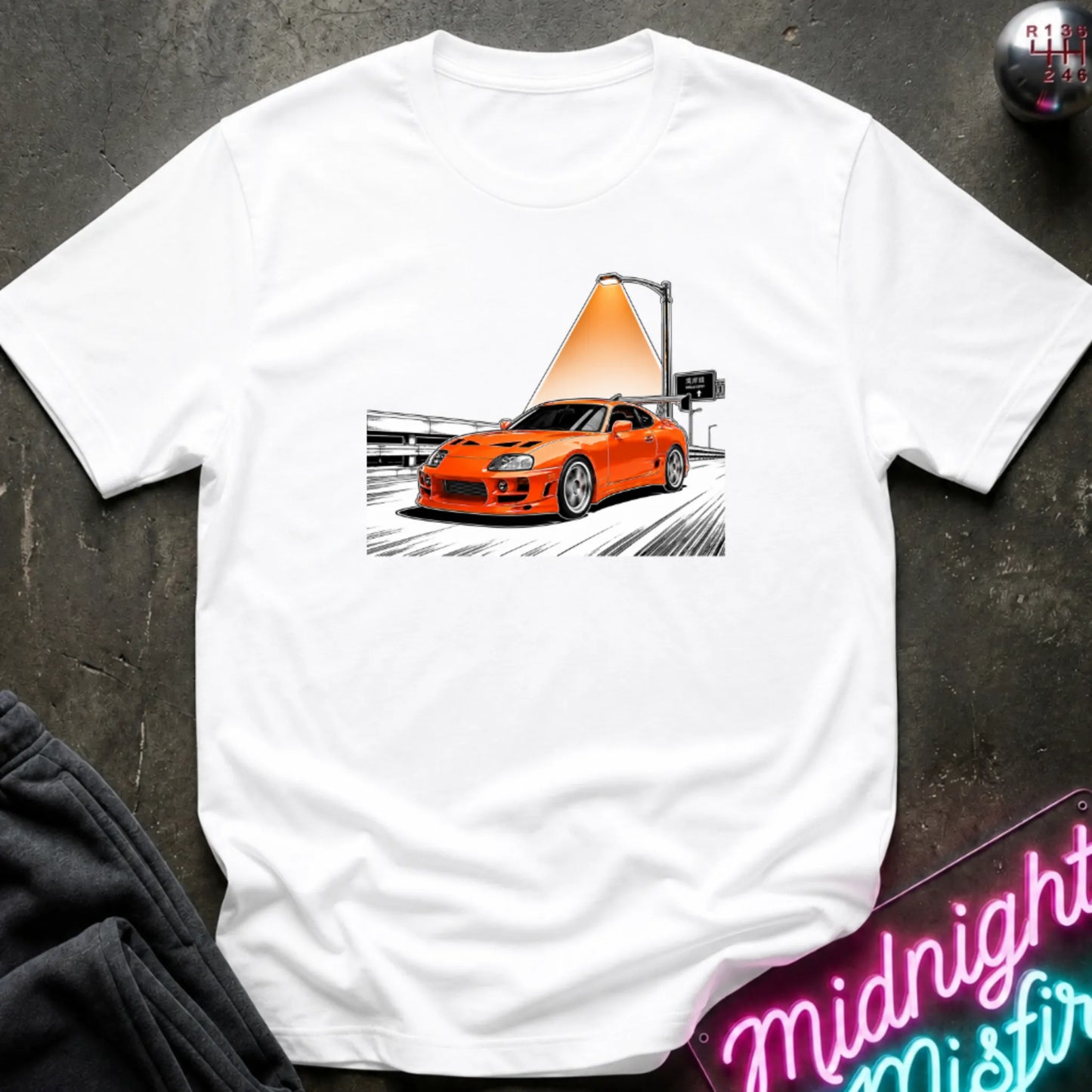 a white  short-sleeve t-shirt laid flat on a dark textured surface, featuring a graphic print of an orange sports car speeding on highway.