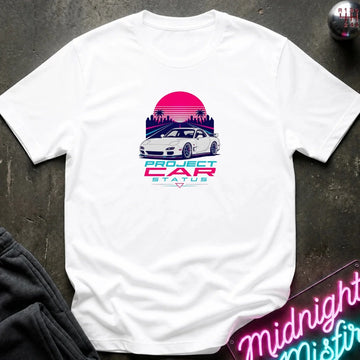 A white short-sleeve t-shirt laid flat on a dark textured surface, featuring a retro 80s-style graphic of a white sports car in front of a pink and purple sunset with the text 'PROJECT CAR STATUS'.