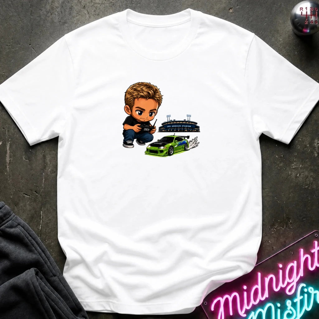 A white graphic t-shirt laid flat on a dark textured surface. The graphic features a green car, and person  controlling it with remote.