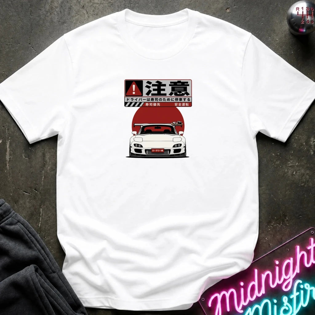 A white short-sleeve t-shirt laid flat on a dark textured surface, featuring a graphic print of a white sports car with a red hood and Japanese text above it. Partially visible props include dark clothing in the lower left and a neon-style sign in the lower right.