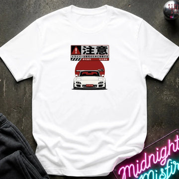 A white short-sleeve t-shirt laid flat on a dark textured surface, featuring a graphic print of a white sports car with a red hood and Japanese text above it. Partially visible props include dark clothing in the lower left and a neon-style sign in the lower right.