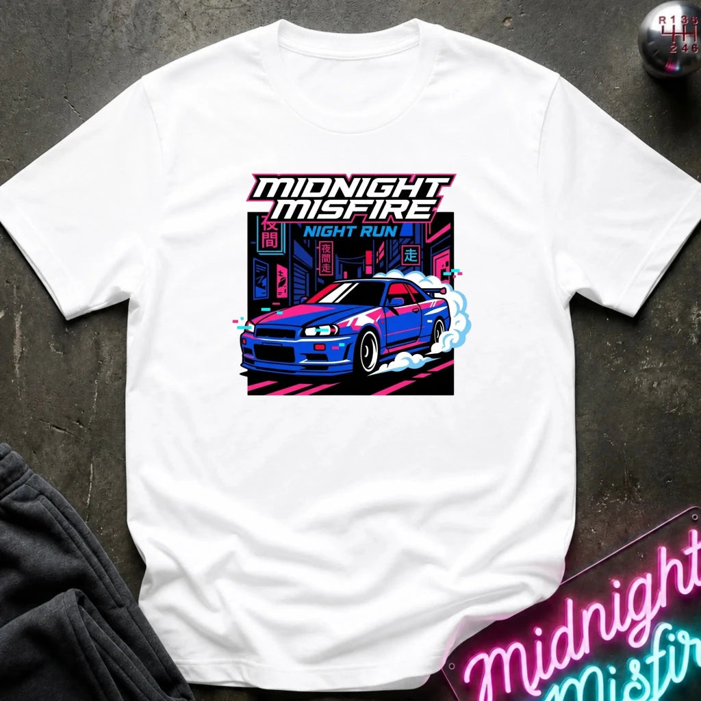 A white short-sleeve t-shirt laid flat on a dark textured surface, featuring a colorful graphic print of a blue sports car in a neon city scene with the text 'MIDNIGHT MISFIRE' and 'NIGHT RUN'.