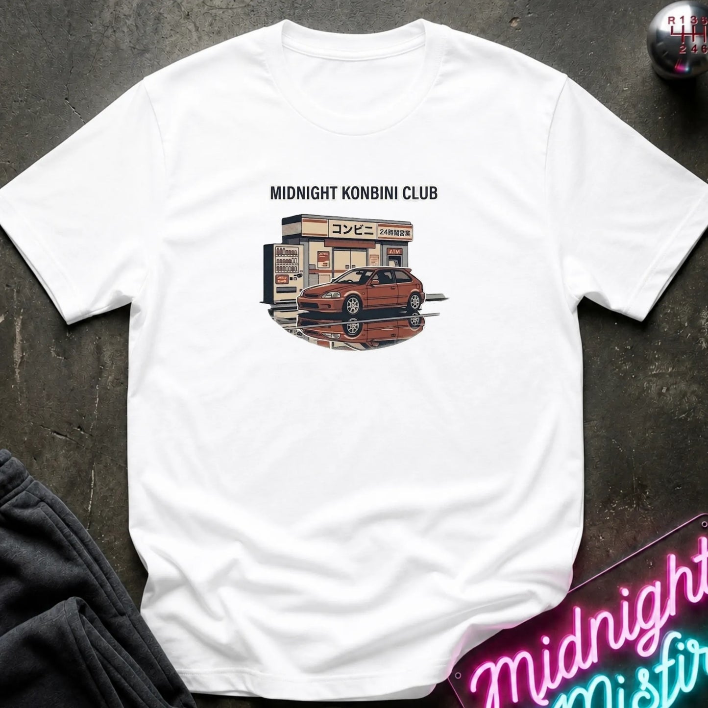 A white short-sleeve t-shirt laid flat on a dark textured surface, featuring a graphic print of a red sports car parked in front of a convenience store with the text 'MIDNIGHT KONBINI CLUB' above it. A neon sign and dark clothing are partially visible as props around the shirt.