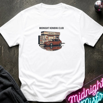 A white short-sleeve t-shirt laid flat on a dark textured surface, featuring a graphic print of a red sports car parked in front of a convenience store with the text 'MIDNIGHT KONBINI CLUB' above it. A neon sign and dark clothing are partially visible as props around the shirt.