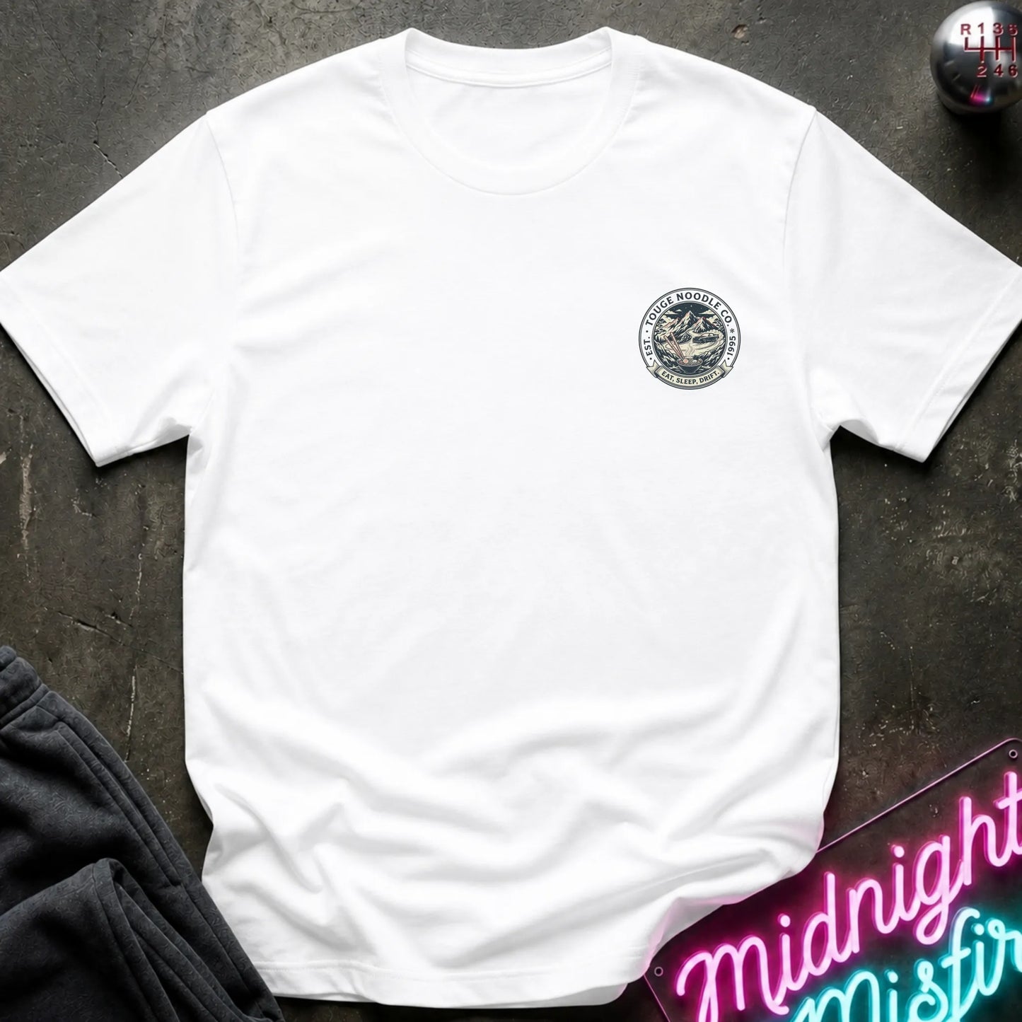 White crewneck t-shirt with a circular crest printed on the left chest, styled flat on a dark textured surface with a neon sign and dark pants in the background.
