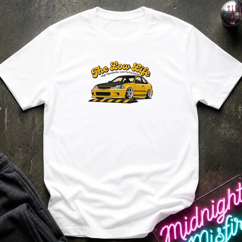 white  t-shirt laid flat on a dark textured surface, featuring a graphic of a yellow sports car with the text 'The Low Life' in yellow script. 