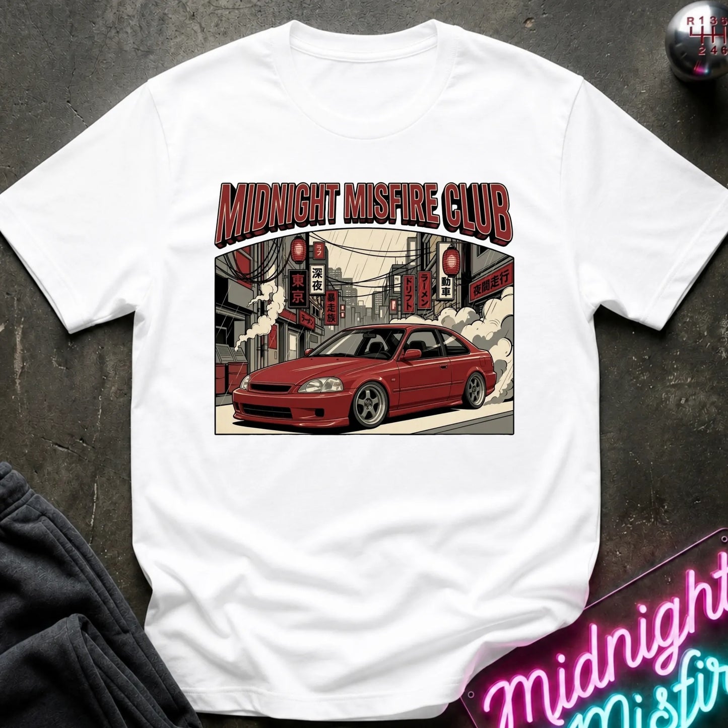A white short-sleeve graphic t-shirt laid flat on a dark textured surface. The graphic features the text 'MIDNIGHT MISFIRE CLUB' above an illustration of a red coupe car driving through a city street with red signs and smoke.