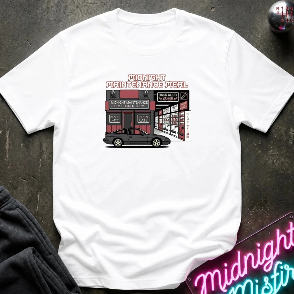 A white short-sleeve t-shirt laid flat on a dark textured surface, featuring a retro-style graphic of a car repair shop with the text 'Midnight Maintenance Meel' and a black car illustration. A neon sign and dark clothing are partially visible in the corners as styling props.