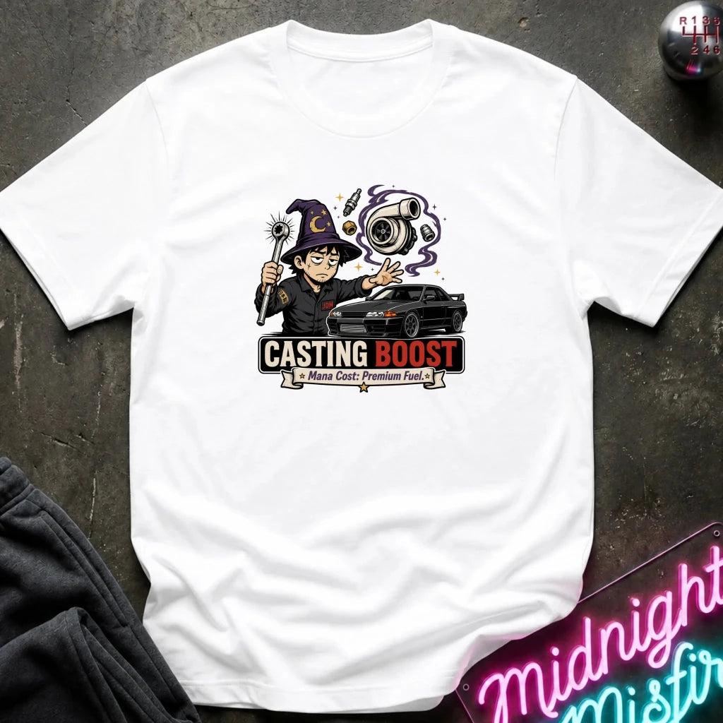 A white short-sleeve graphic T-shirt laid flat on a dark textured surface. The shirt features a colorful cartoon wizard holding a wand next to a black car, with the text 'CASTING BOOST' and smaller text 'Majestic Premium Fuel.