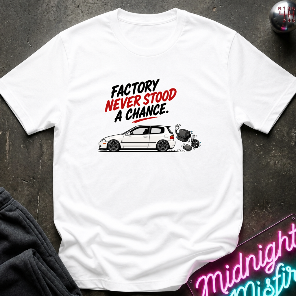 A white short-sleeve graphic t-shirt laid flat on a dark textured surface. The graphic features the text 'FACTORY NEVER STOOD A CHANCE.