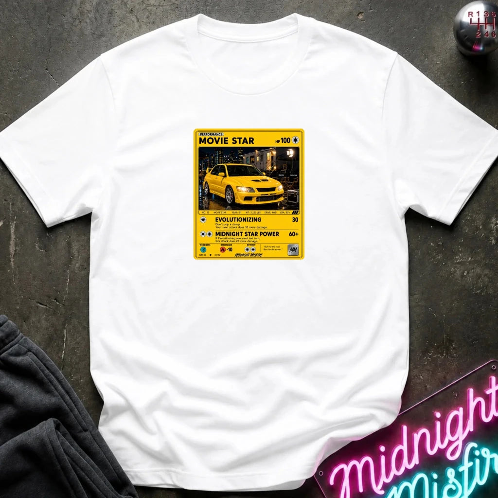 white short-sleeve t-shirt laid flat on a dark textured surface, featuring a yellow and black retro-style graphic on the chest with a yellow car photo and text including 'MOVIE STAR'. 