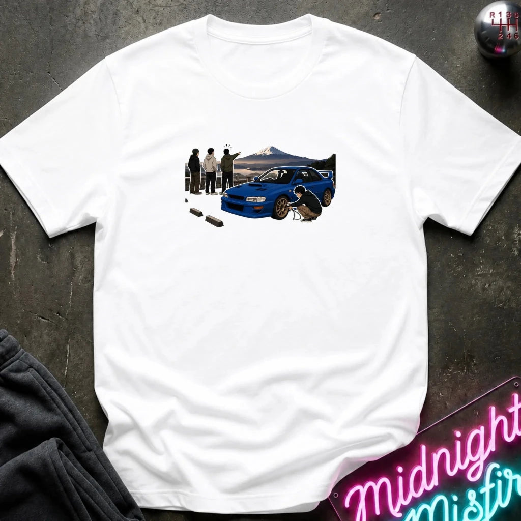 white crewneck t-shirt laid flat on a dark textured surface. The shirt features a graphic print of a blue sports car with a driver and spectators in front of a mountain landscape.
