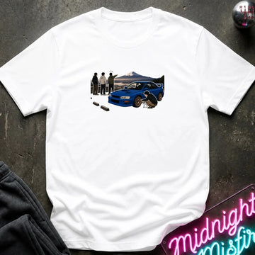 white crewneck t-shirt laid flat on a dark textured surface. The shirt features a graphic print of a blue sports car with a driver and spectators in front of a mountain landscape.